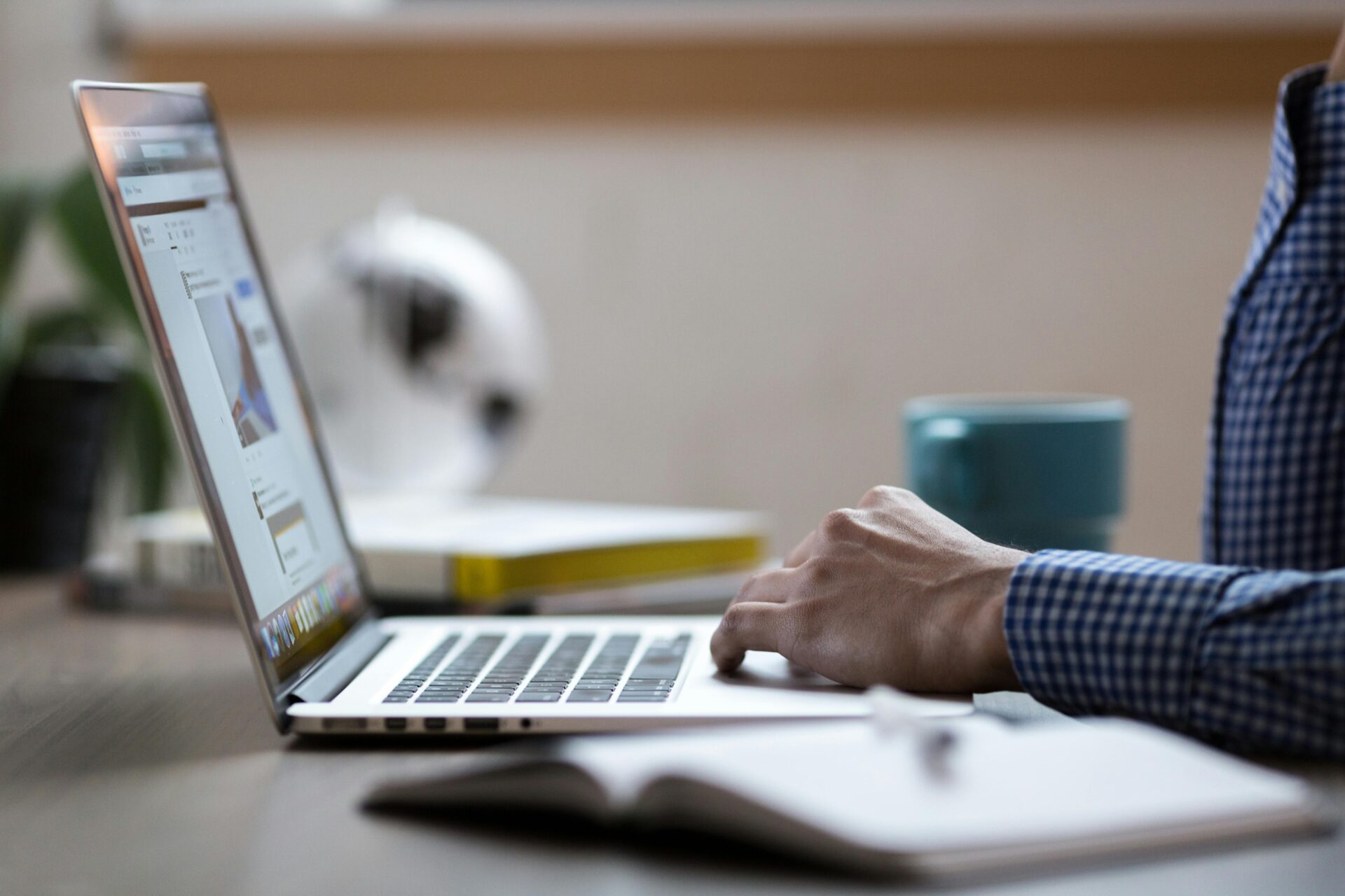 Side view of a person’s hand using a laptop trackpad on a wooden desk with a notebook, coffee mug, and globe in the background.