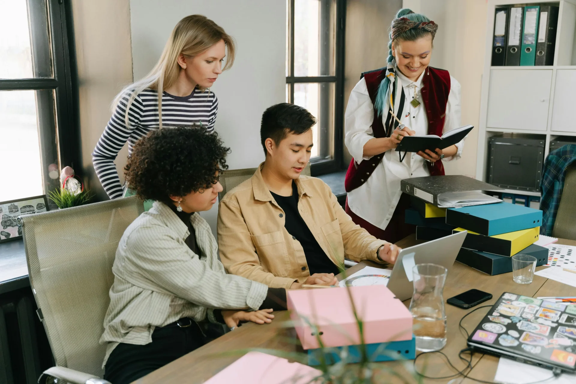 A diverse team of four colleagues gathered around a laptop in an office discussing the benefits of video in marketing.