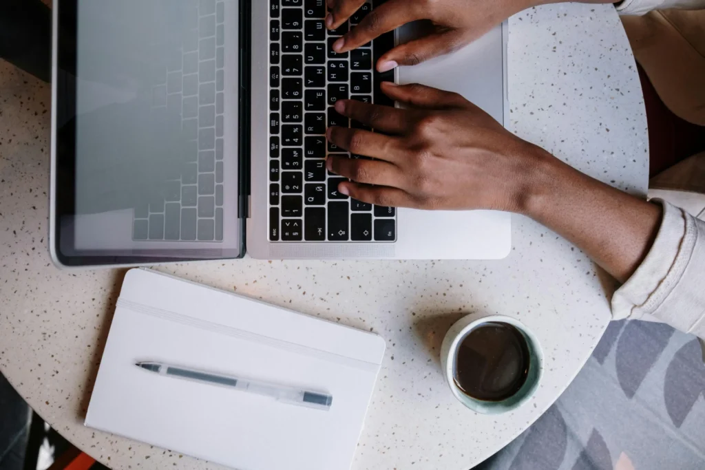 A person typing on a laptop keyboard next to a white notebook and a cup of coffee on a speckled table, representing contact page essentials for modern businesses.