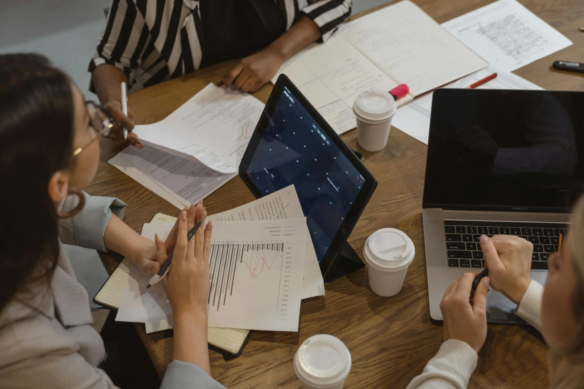 Four colleagues sitting around a wooden table with a tablet, laptop, and coffee cups, reviewing printed financial reports and digital calendars.