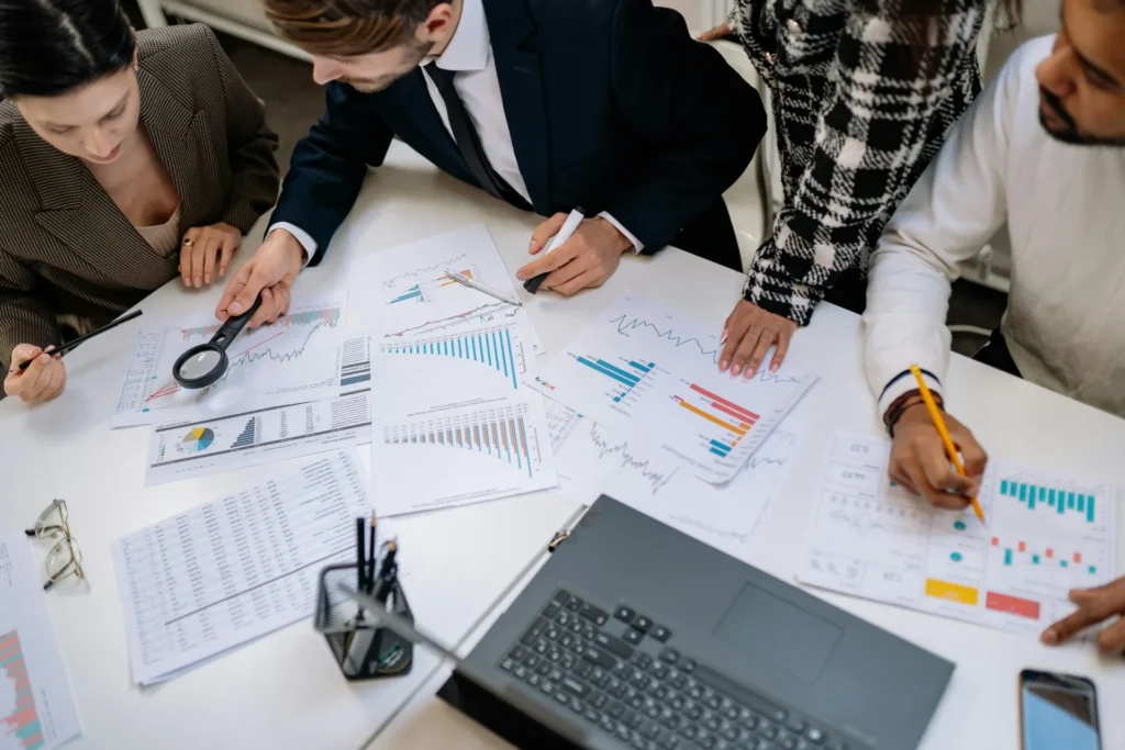 A business team at a white desk using a magnifying glass to review printed data charts and graphs for a digital marketing campaign.