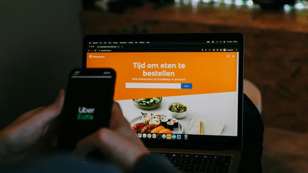 Person using laptop and smartphone for online food ordering via Uber Eats, illustrating digital marketing for food and beverage services.