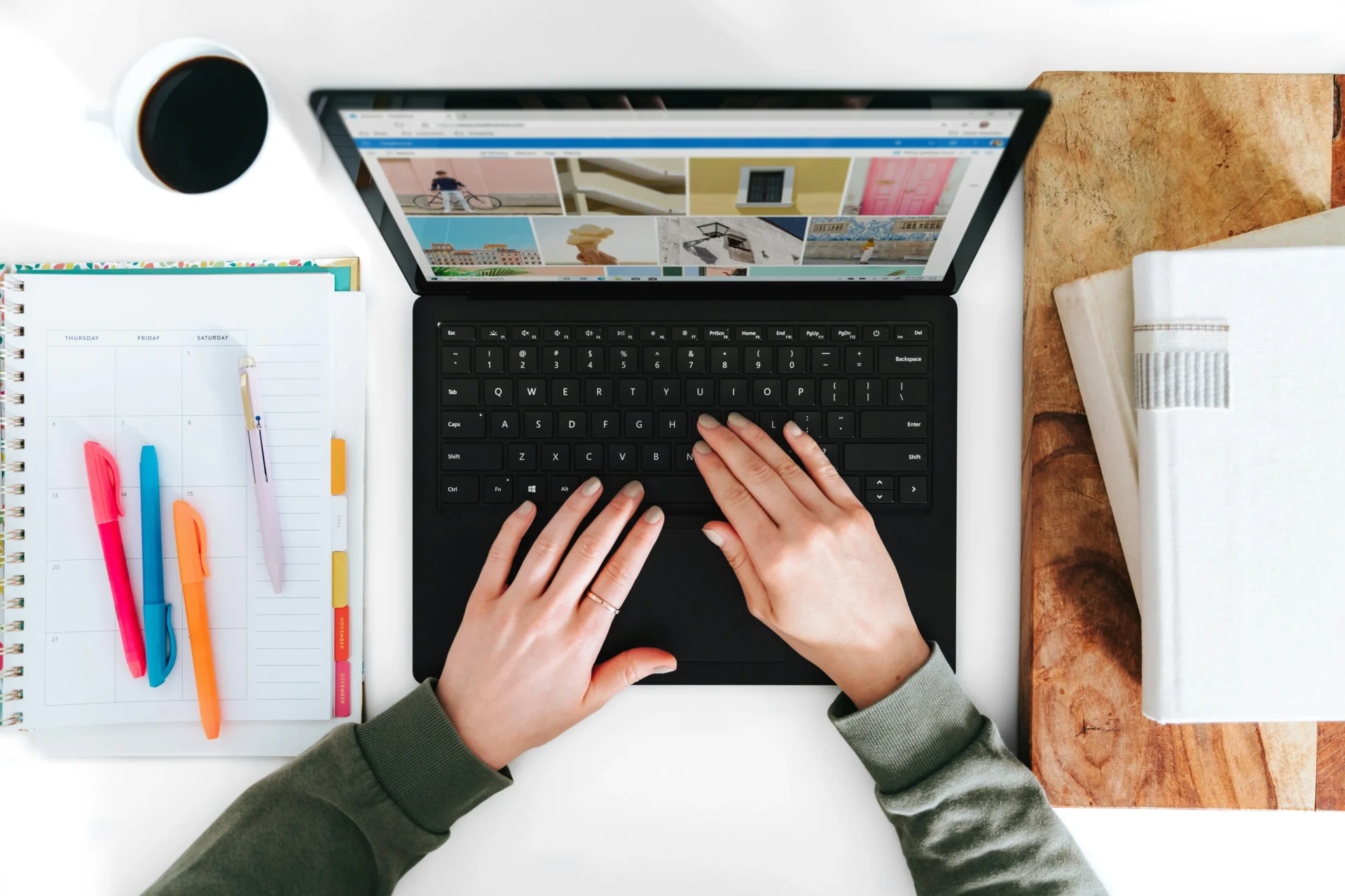 Hands typing on a laptop keyboard, working on SEO for food and beverage content, with a notebook, pens, coffee, and books on a white desk.