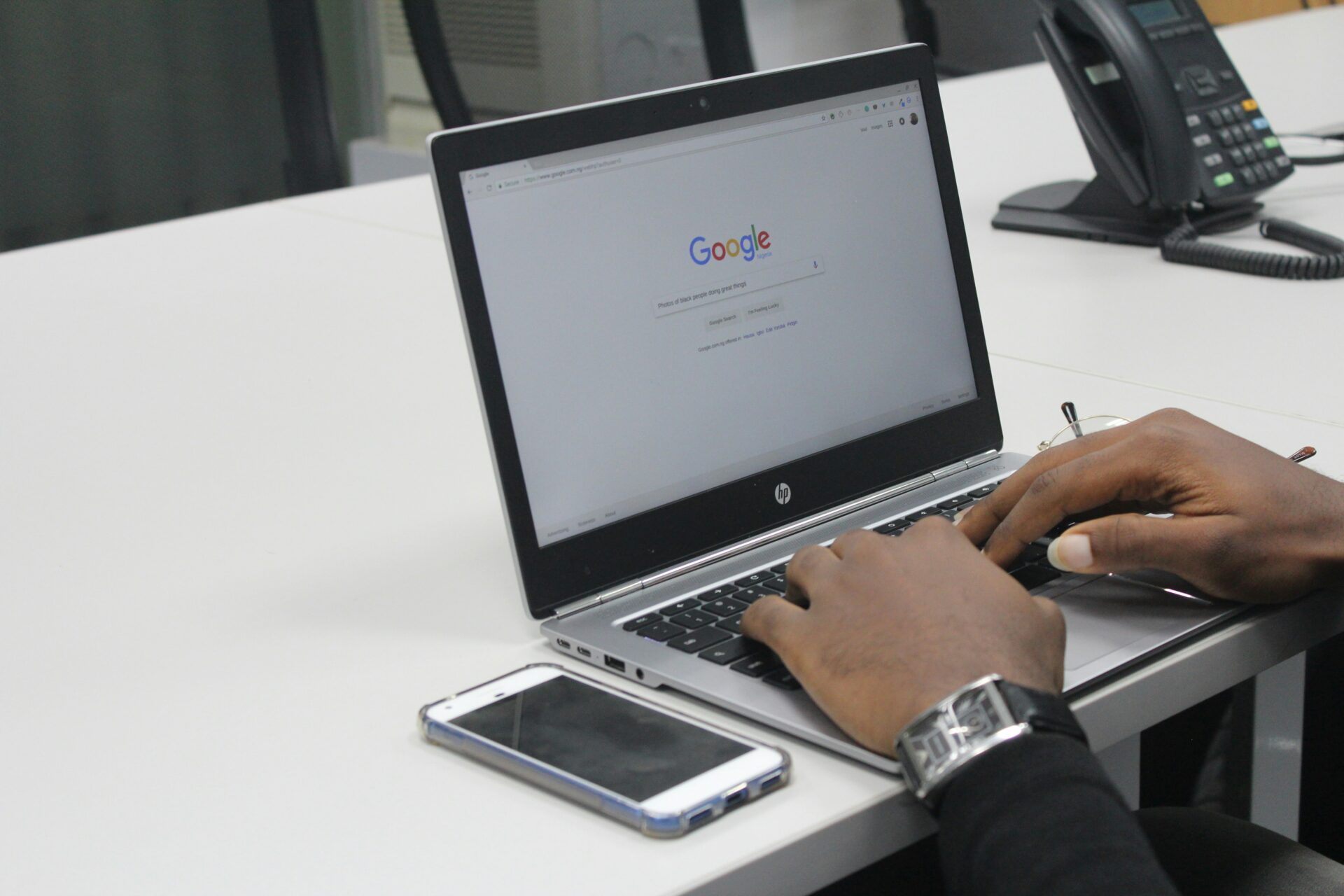 Hands typing on a laptop with Google search page visible, smartphone and office phone nearby.