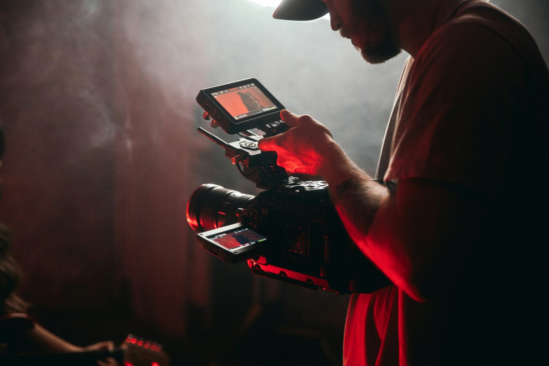 Filmmaker operating a professional camera with an external monitor in a smoky, red-lit environment.