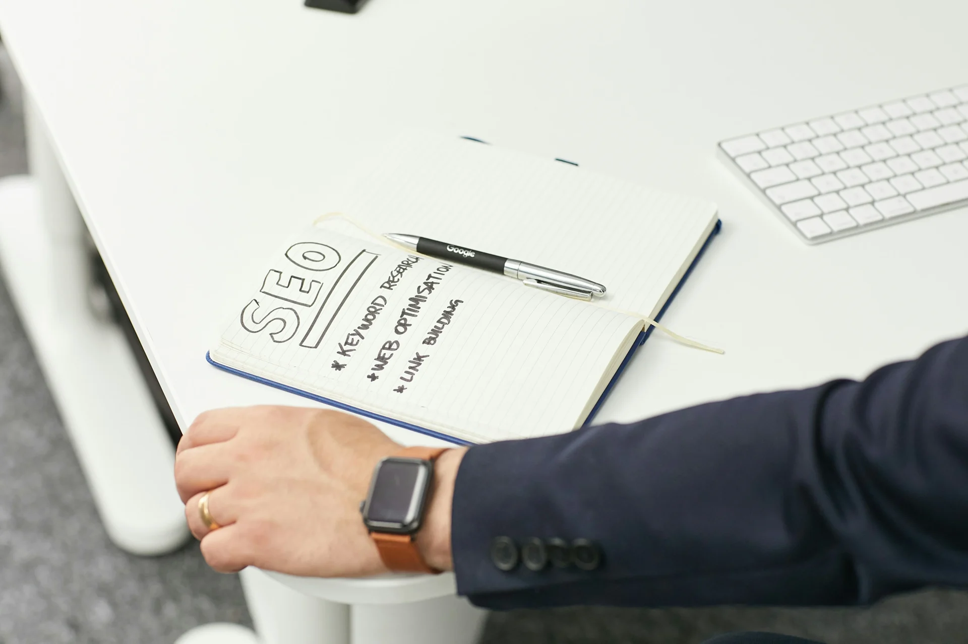 Close-up of a notebook with "SEO" handwritten, listing keyword research, web optimisation, and link building, next to a pen and a person's arm wearing a smartwatch, implying the creation of an on-page SEO checklist.