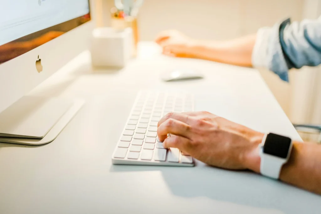 Person's hands typing on a white keyboard and using a mouse at a clean desk with an Apple monitor.