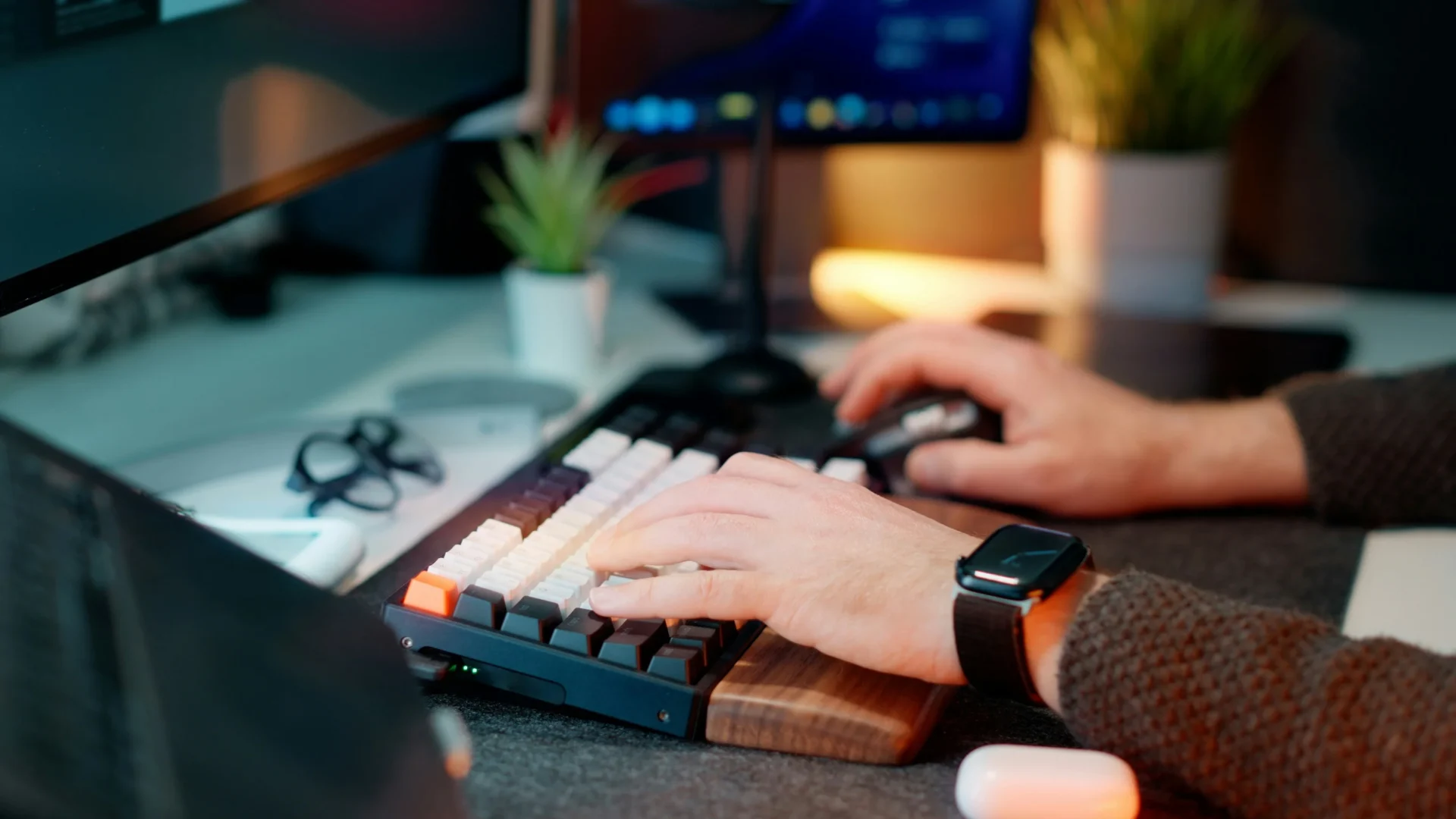 Close-up of hands typing on a mechanical keyboard with a wooden wrist rest and using a mouse, showing a home office setup.
