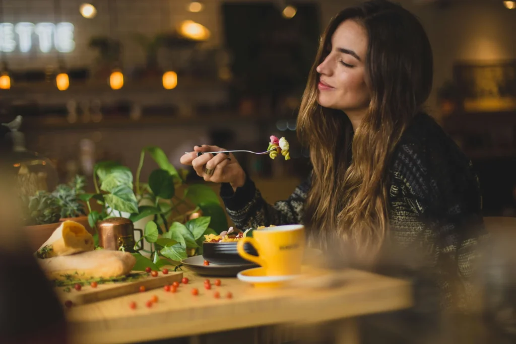 A woman with long hair enjoying a meal in a restaurant, holding a fork with food, with a yellow cup and plants on the table.