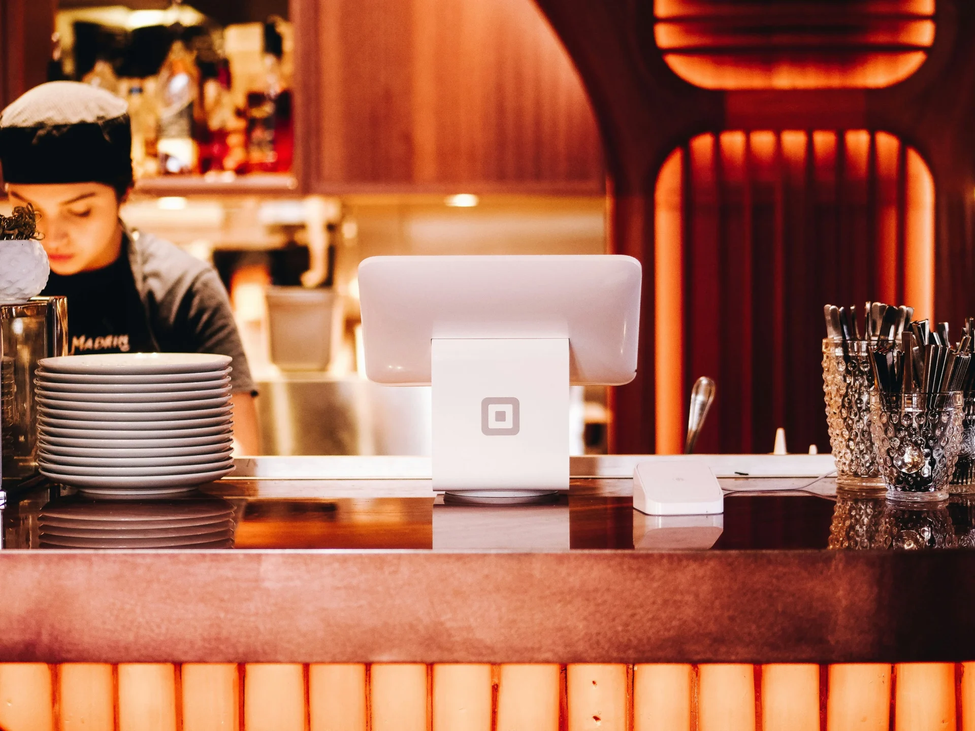 A Square point-of-sale system on a restaurant counter with a stack of plates and a person working in the background.