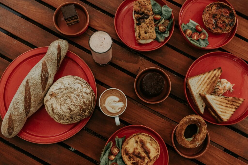 An overhead shot of various food and beverage items including bread, pastries, sandwiches, and coffee on a wooden table, illustrating a comprehensive food and beverage marketing plan.