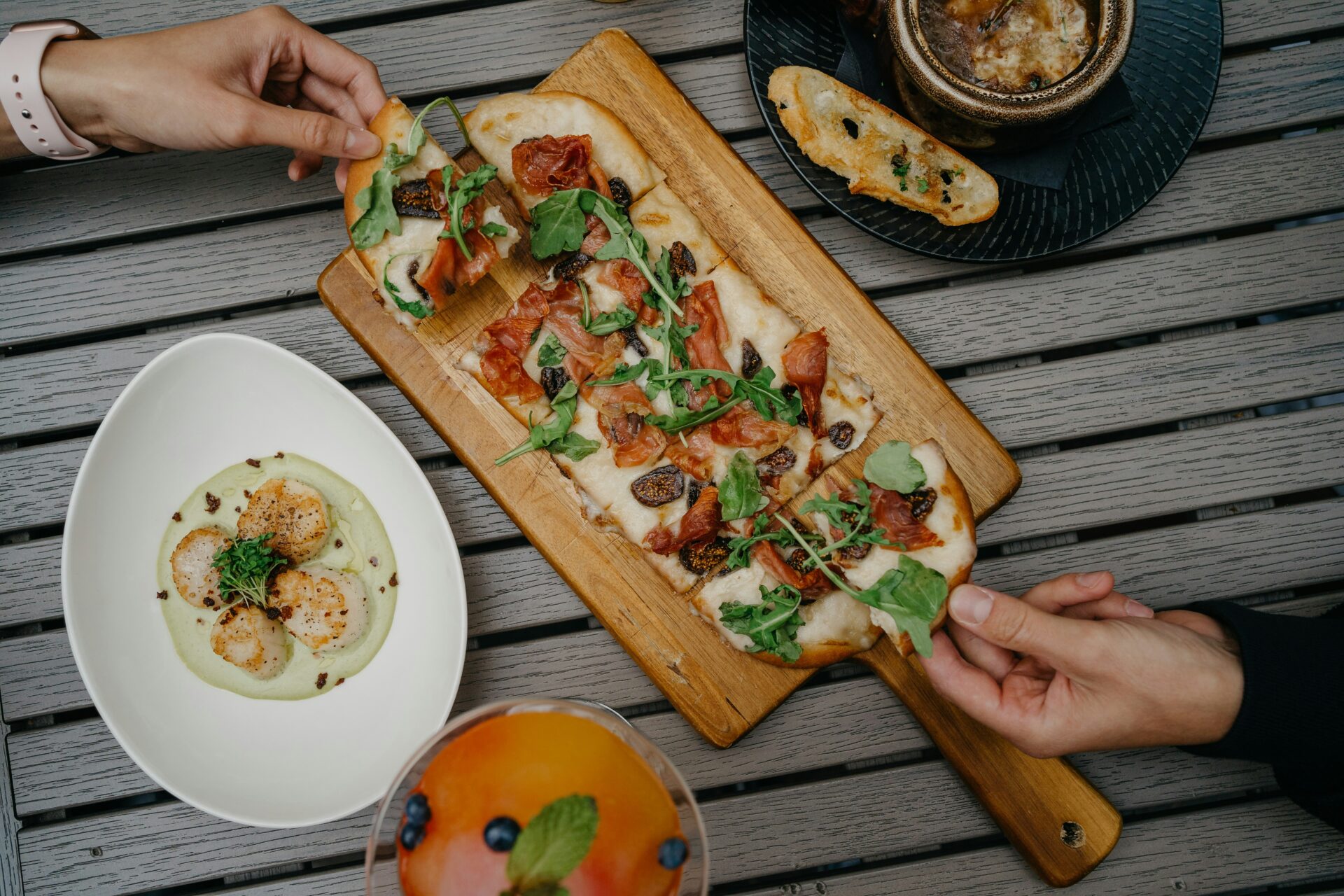 Hands reaching for slices of flatbread pizza on a wooden board, with scallops and a drink on a table, representing a food and beverage marketing plan.