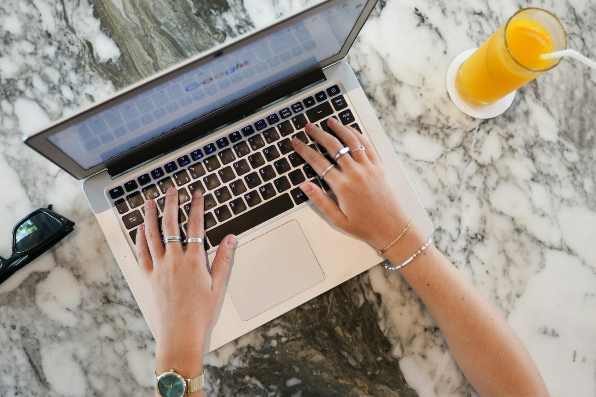 Hands typing on a laptop keyboard with Google open on the screen, an orange juice, and sunglasses on a marble table, representing national search engine optimization efforts.