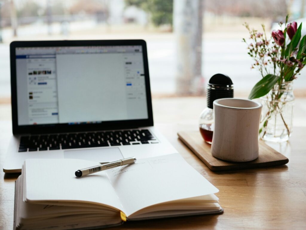 Laptop and open notebook with a pen on a wooden table, with a coffee cup and flowers in the background, perfect for a content marketing brief.