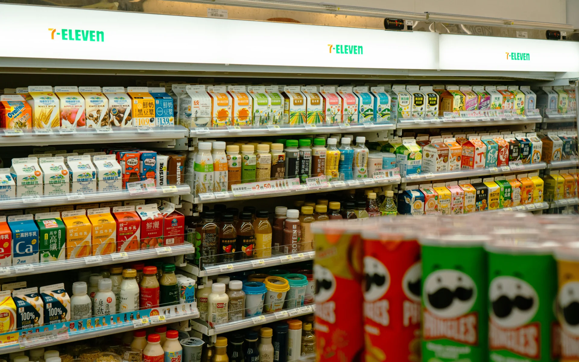 Aisle in a 7-Eleven convenience store filled with various CPG beverage products like milk, juice, bottled drinks, and Pringles cans.
