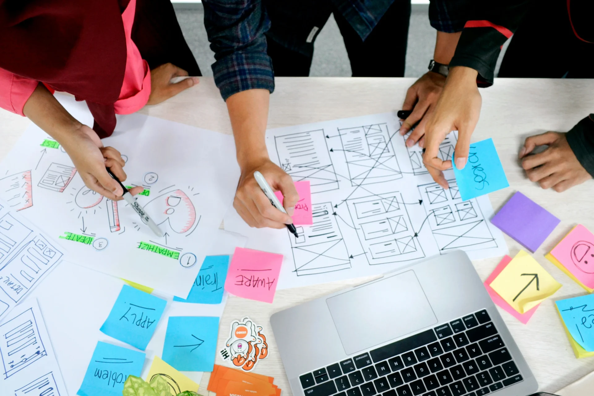Top-down view of a team collaborating on a design project with diagrams, sticky notes, and a laptop on a white table.
