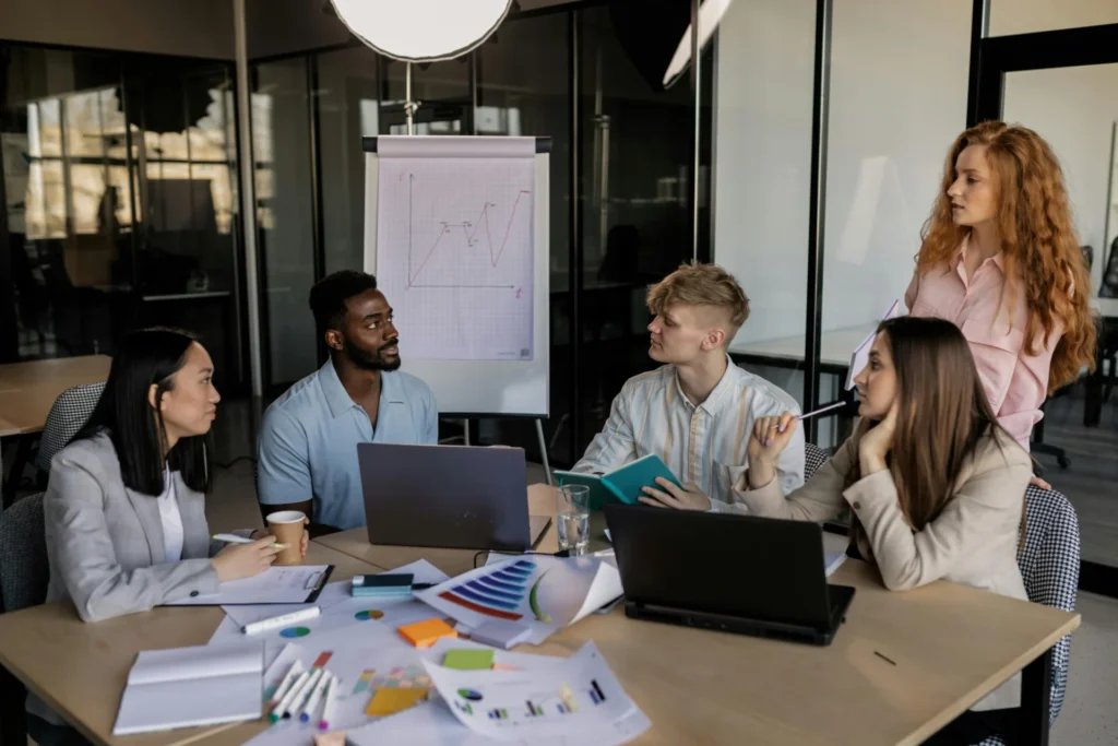 A diverse team of five people collaborates around a table with laptops and charts, working together on marketing strategies for small businesses.
