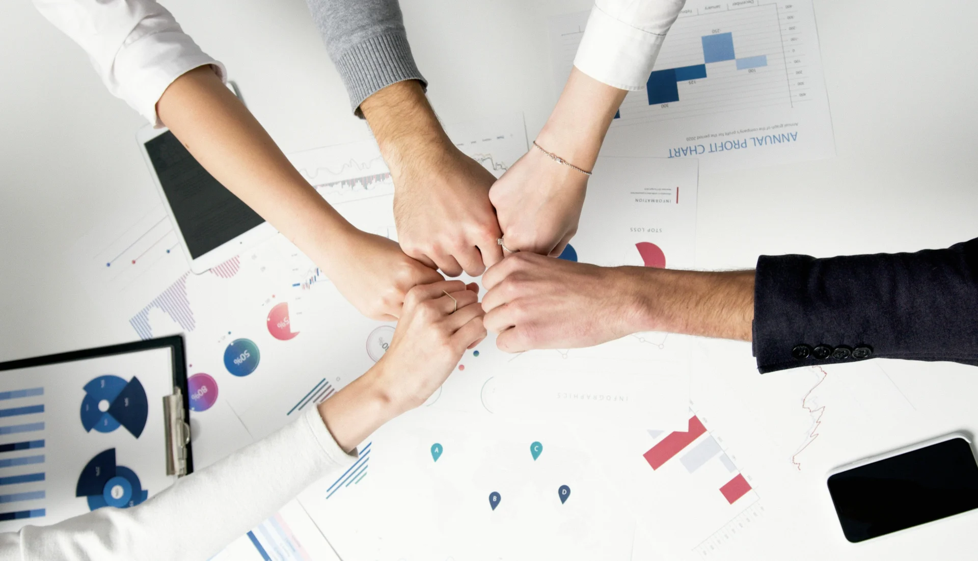 An overhead shot of four people giving a celebratory fist bump over a table covered with business charts and graphs.