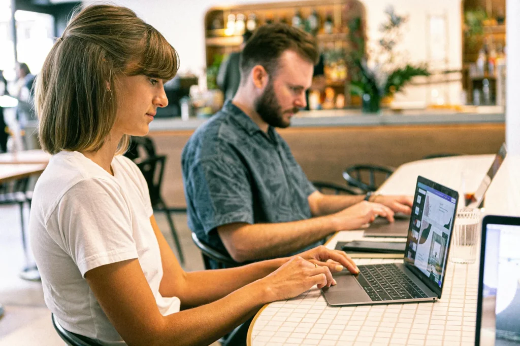 A man and a woman are seated at a long table in a cafe, both focused on their laptops, with one laptop screen showing a website design.