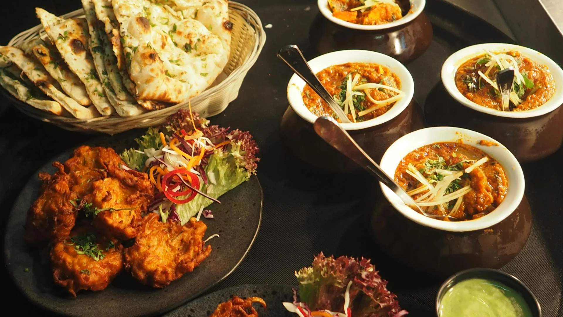 A close-up shot of a tray of assorted Indian dishes, including naan bread in a basket, pakoras on a plate with salad, and several bowls of curry.