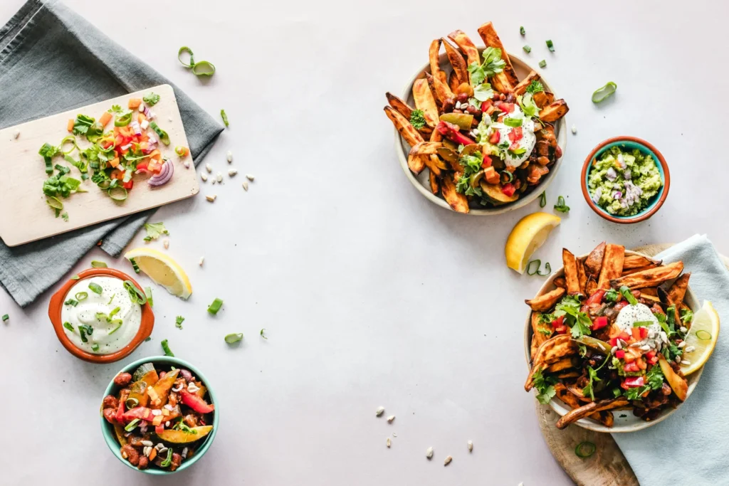 A top-down view of two bowls of loaded sweet potato fries with various toppings, surrounded by small bowls of salsa, guacamole, and a cutting board with chopped vegetables.