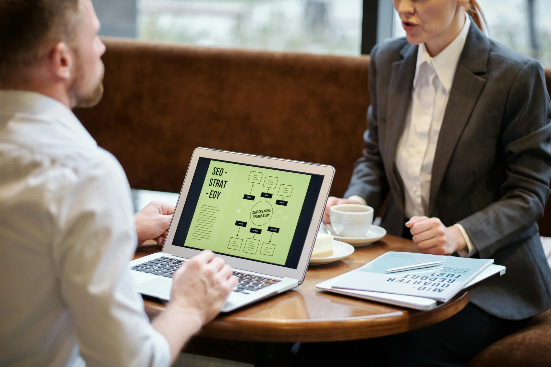 Two professionals are sitting at a cafe table, with a laptop displaying a mind map about SEO strategy, and a report on the table.