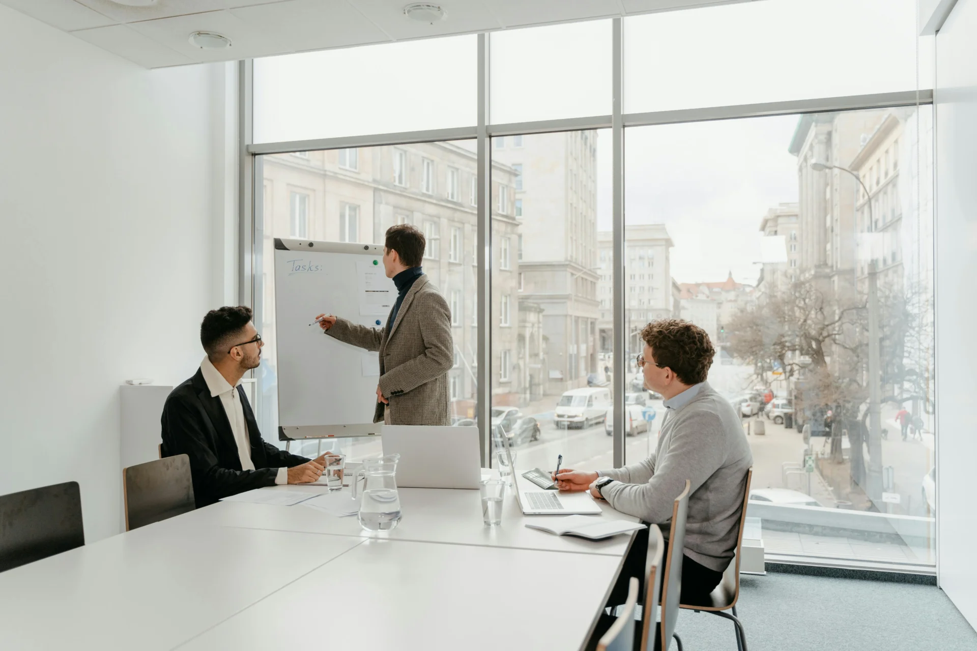 Three professionals in a bright meeting room with large windows discussing how can i get leads for my business, using a whiteboard to outline a strategy.