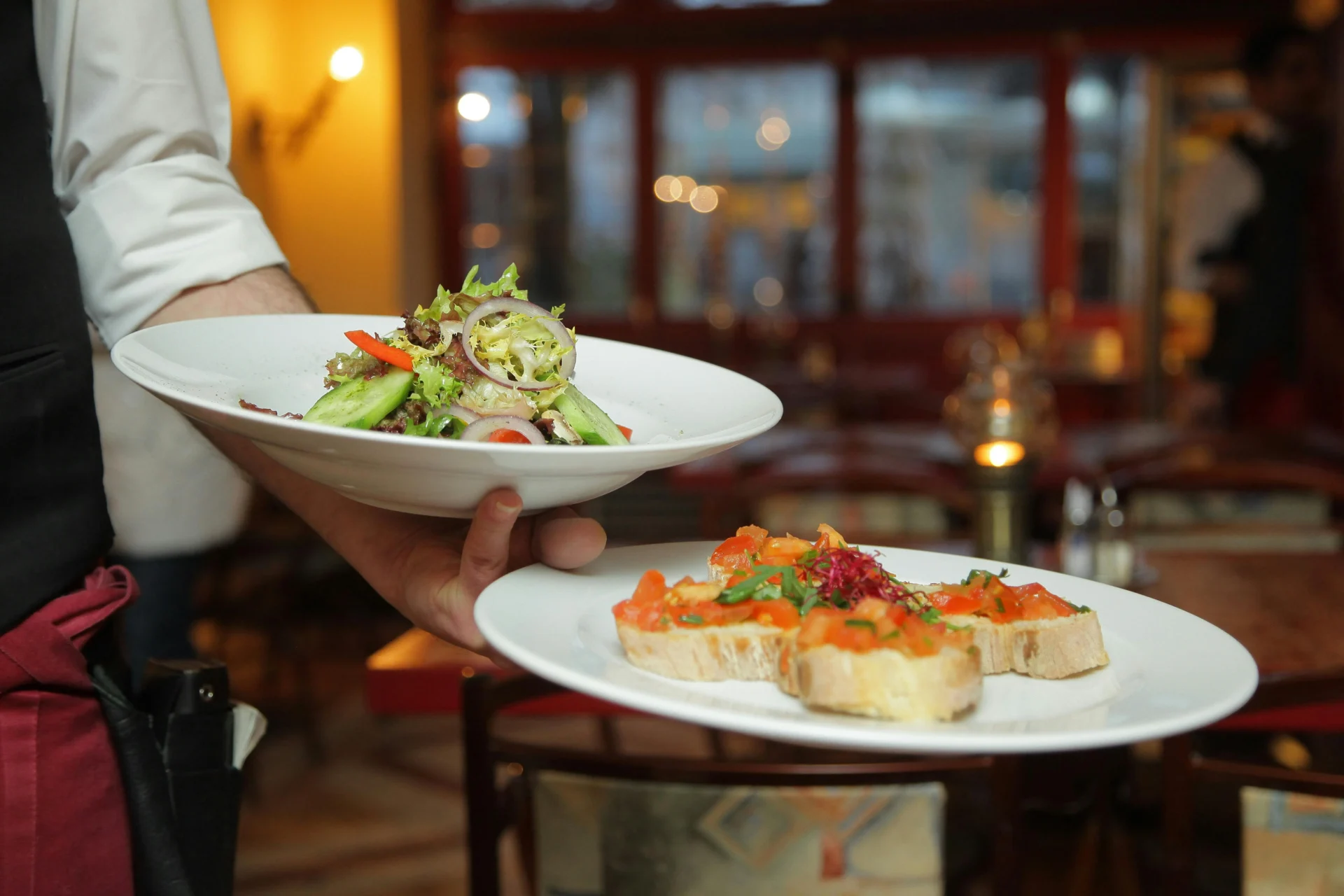 A waiter in a restaurant is holding two plates of food, a green salad, and bruschetta, in a soft-focus setting with warm lighting.