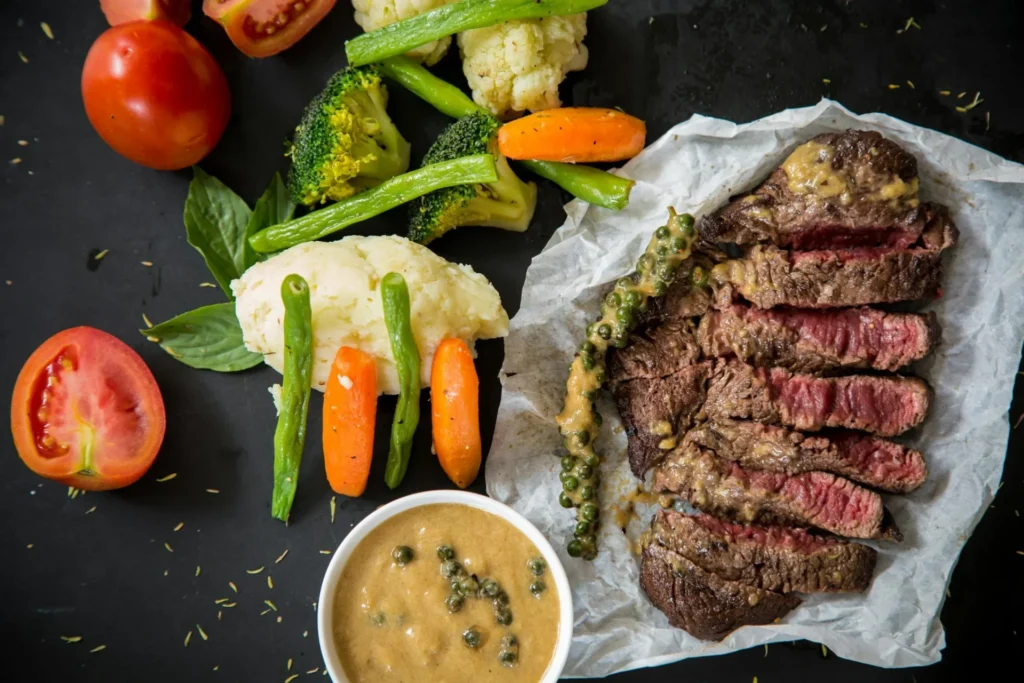 A top-down shot of a perfectly cooked steak, sliced and drizzled with sauce, next to a bowl of sauce and various steamed vegetables like carrots, cauliflower, and broccoli.