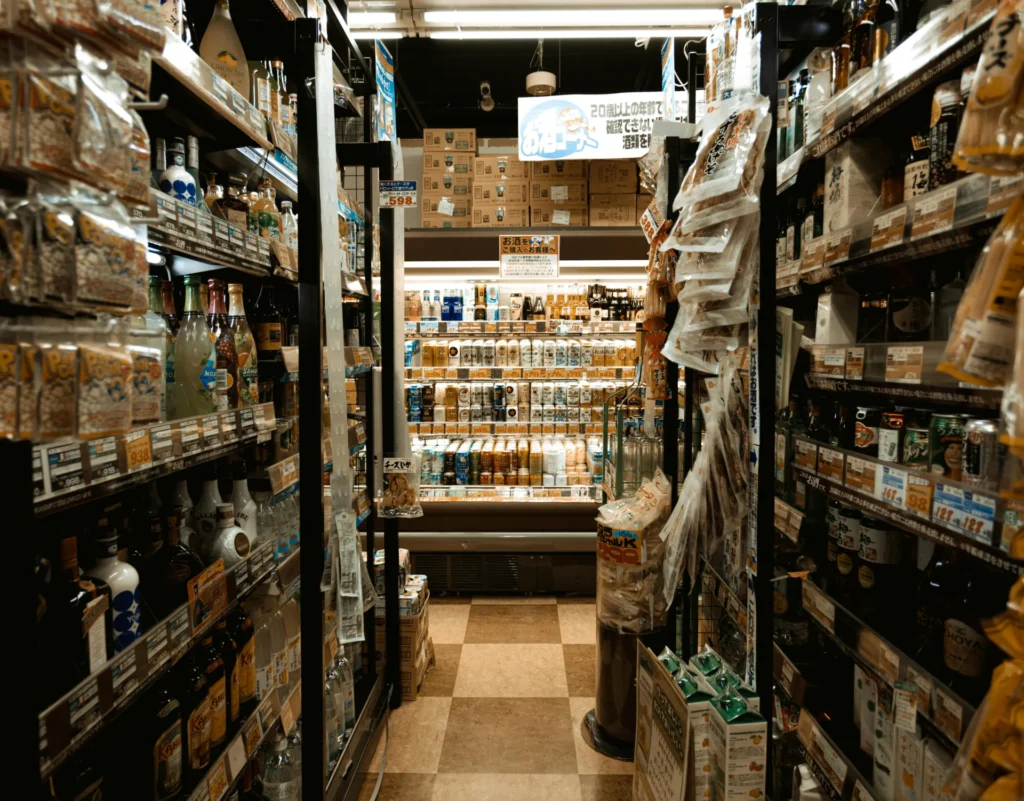 A narrow aisle in a Japanese convenience store with shelves on both sides, leading to a brightly lit refrigerated section with drinks.