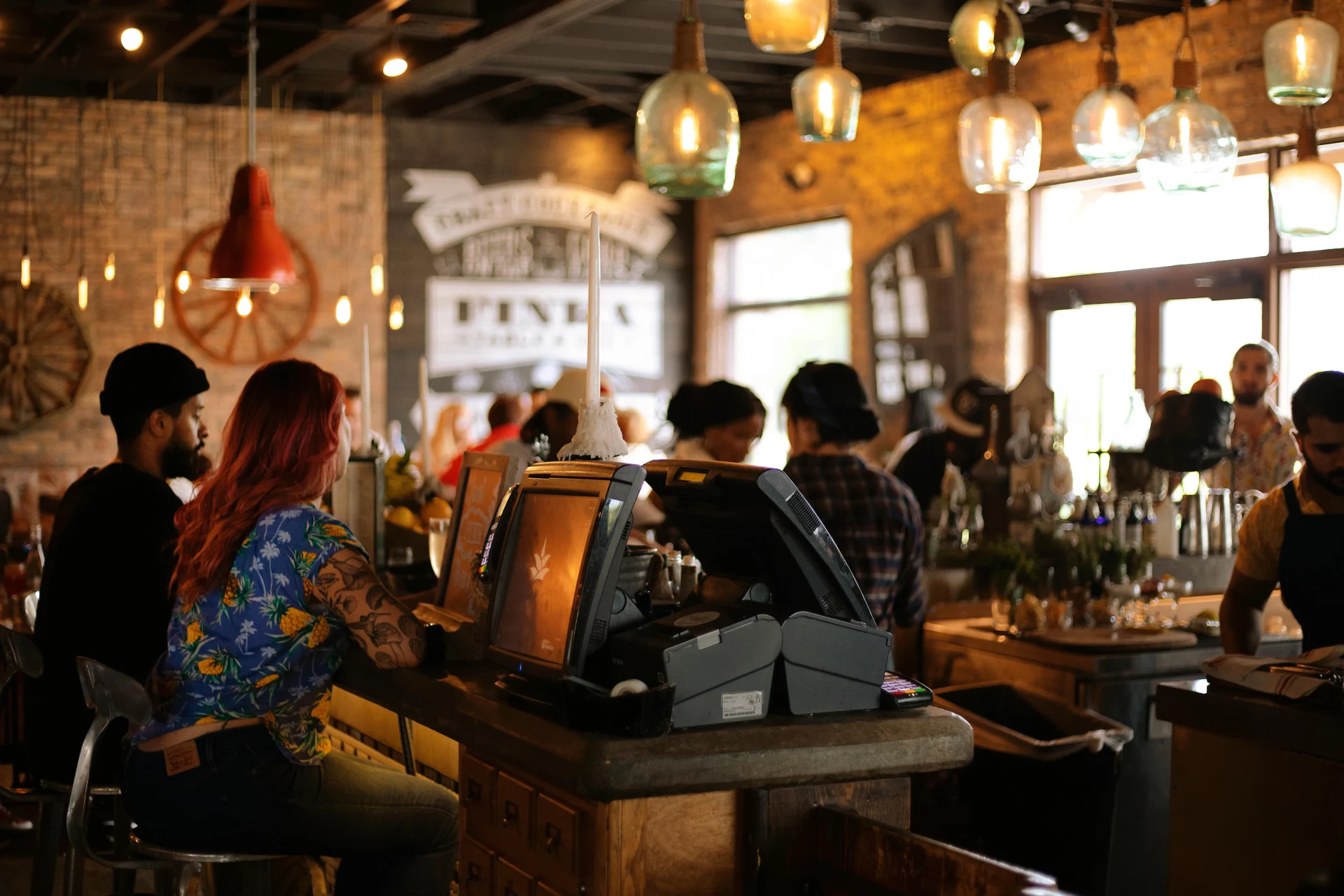 Customers and staff at a busy, dimly lit, trendy bar or restaurant with exposed brick walls, vintage lighting, and a point-of-sale system on the counter.