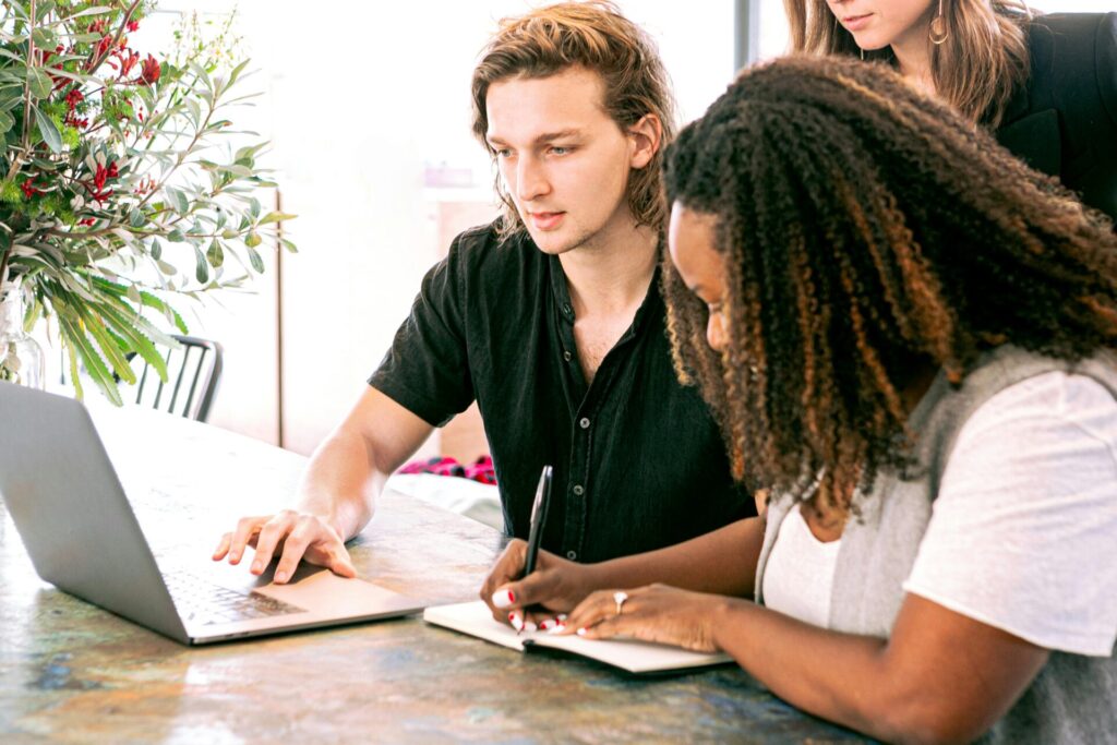 Two people, one with long hair and a black shirt and the other with curly hair, sit together at a table looking at a laptop. The person with curly hair is writing in a notebook, while the person in the black shirt points at the screen. A third person is partially visible in the background.