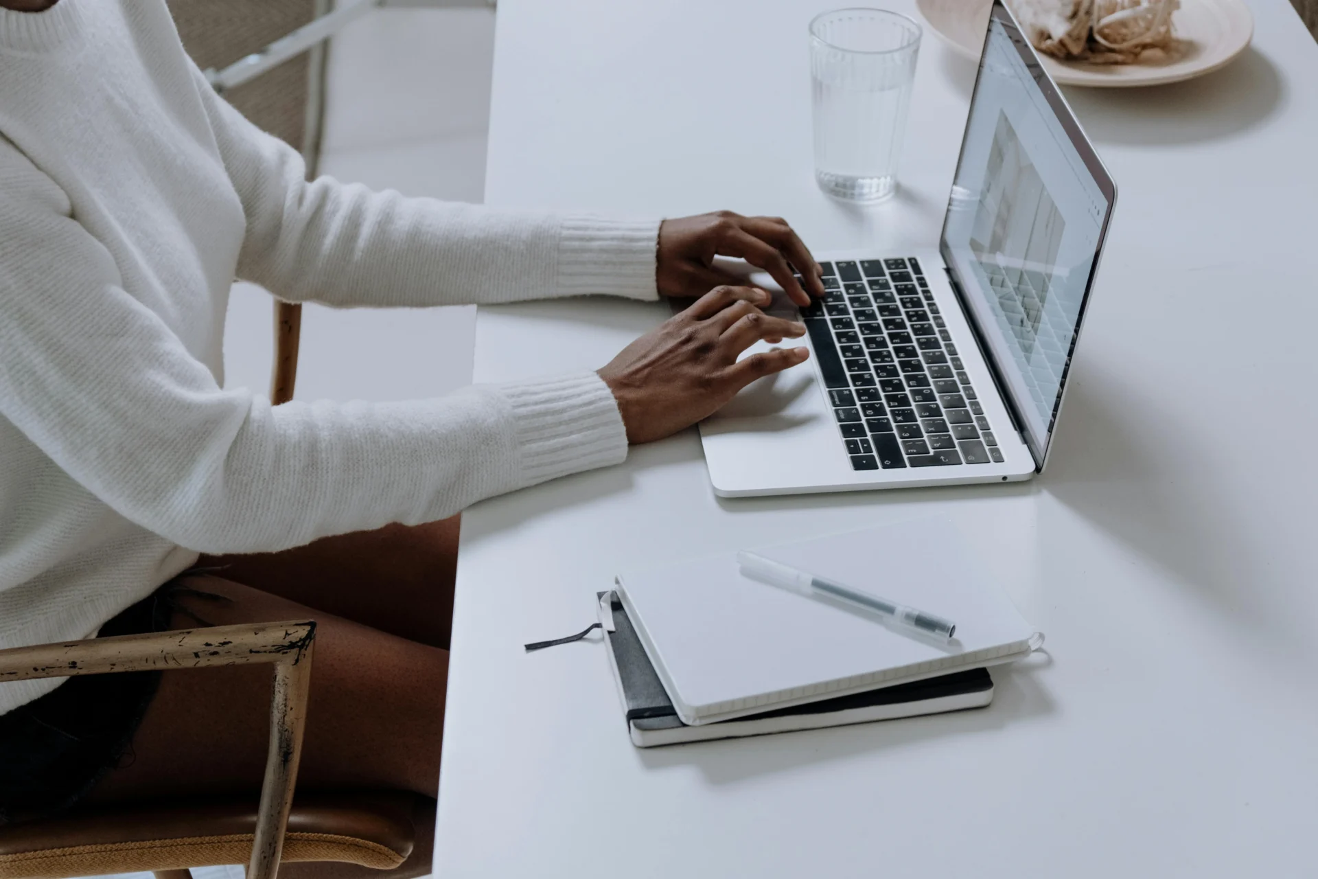 A person wearing a white sweater types on a silver laptop at a white table with two notebooks, a pen, and a glass of water.