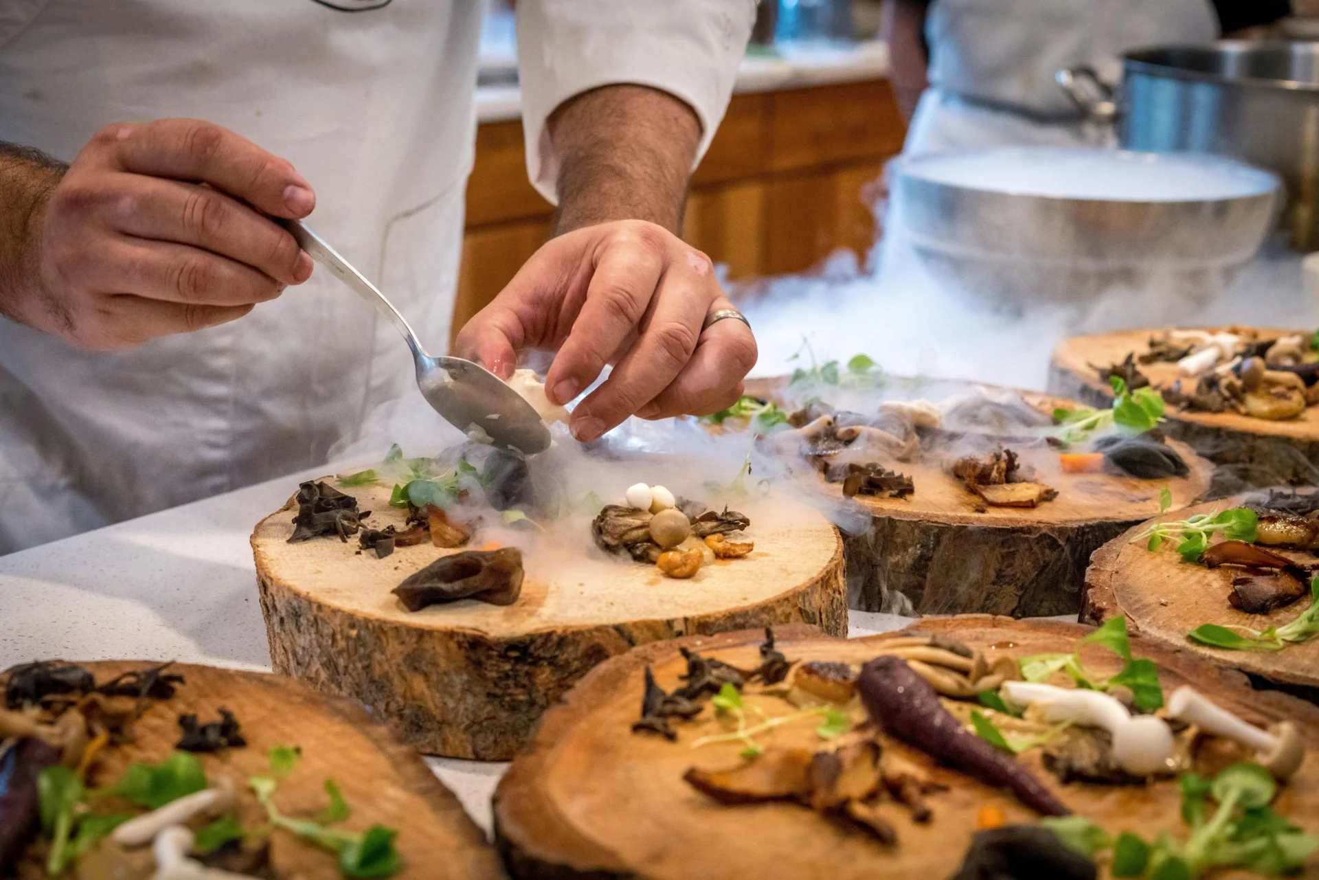 A professional chef in a white coat plates a gourmet dish of sautéed mushrooms and microgreens on a rustic wooden platter, using a spoon to add a final touch. Dry ice creates a smoky, dramatic effect around the plates. The scene highlights the artistry and plating of restaurant cuisine.