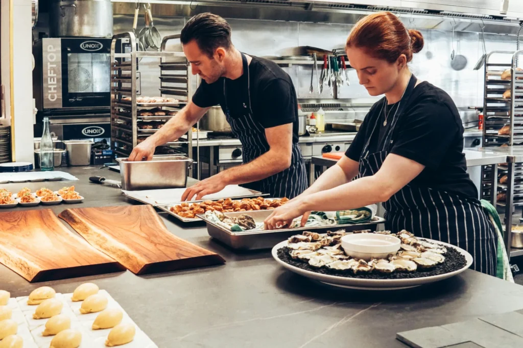 Two chefs in black shirts and striped aprons, a man and a woman, prepare dishes in a stainless steel commercial kitchen.