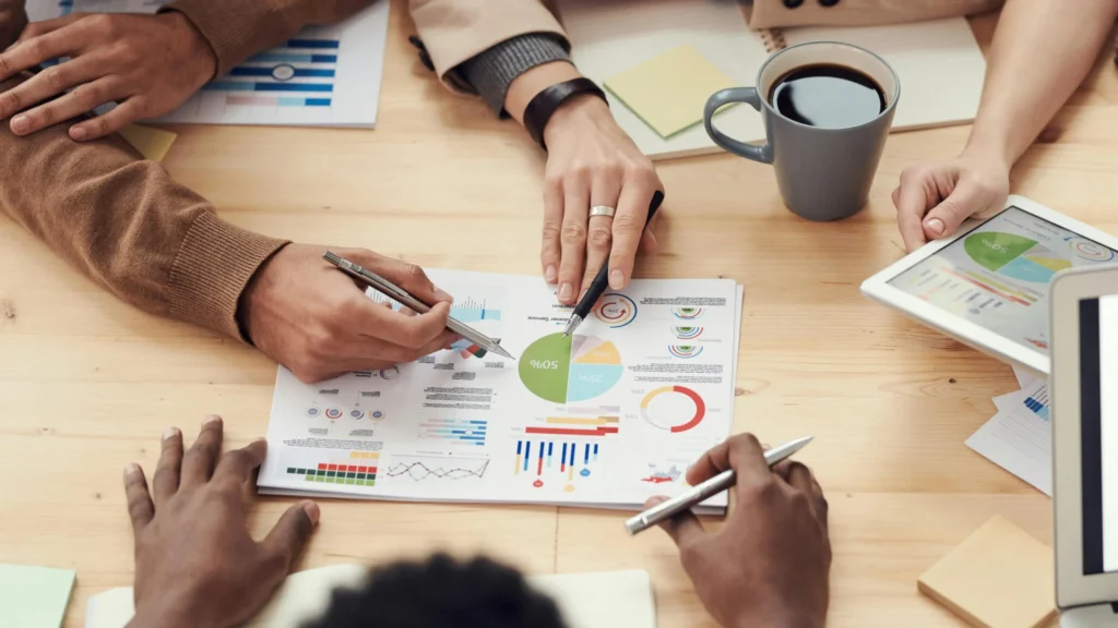 An overhead shot of three people's hands at a wooden table, pointing at a paper with colorful graphs and charts. A laptop, a tablet showing a graph, and a coffee mug are also on the table. The image shows a focused discussion on data analysis or a business report.
