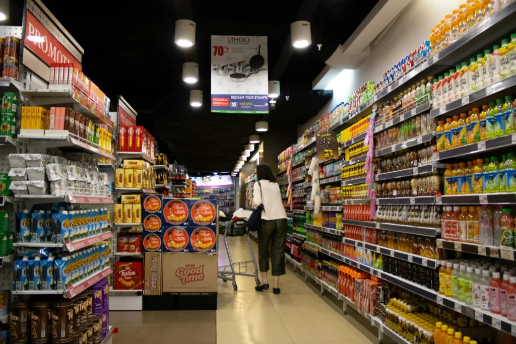 A woman pushes a shopping cart down a grocery store aisle lined with shelves of brightly colored consumer goods, including drinks and packaged food.