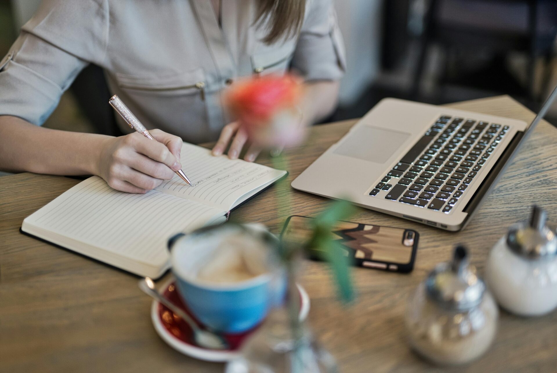 A person sits at a cafe table with a latte and a laptop, writing in a notebook with a pen. The scene suggests a focused moment of creative planning or strategy creation.