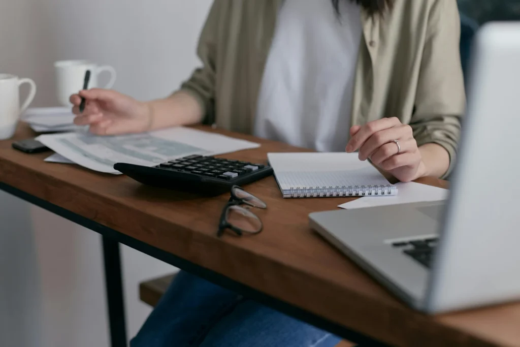 A person sits at a wooden desk with a laptop, calculator, and paperwork, including a blank notepad and two mugs. The person is holding a pen and using the calculator, appearing to be working on finances or business planning.