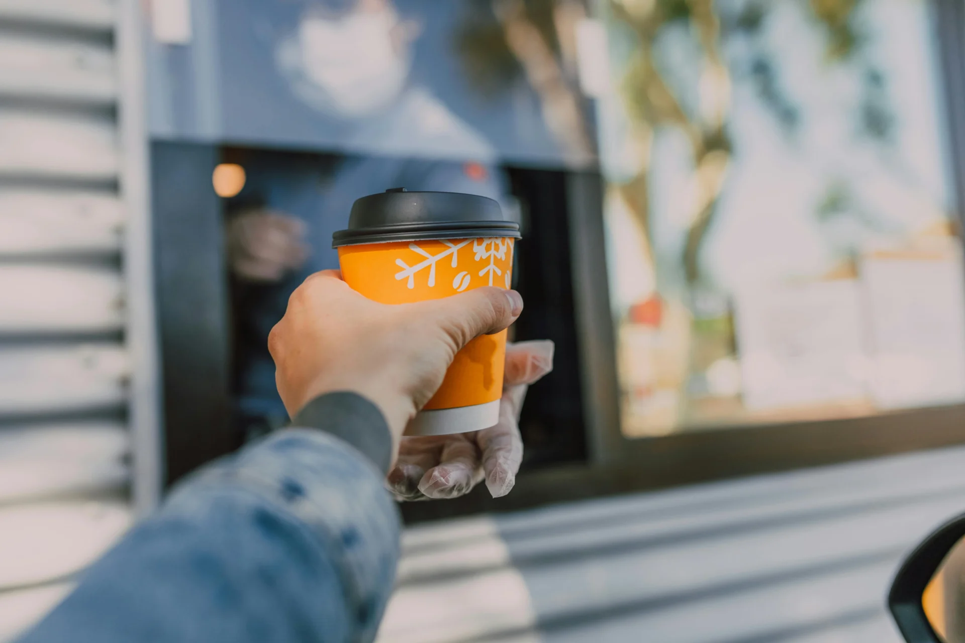 A hand reaches out to receive an orange and white disposable coffee cup from a drive-thru window on a sunny day.