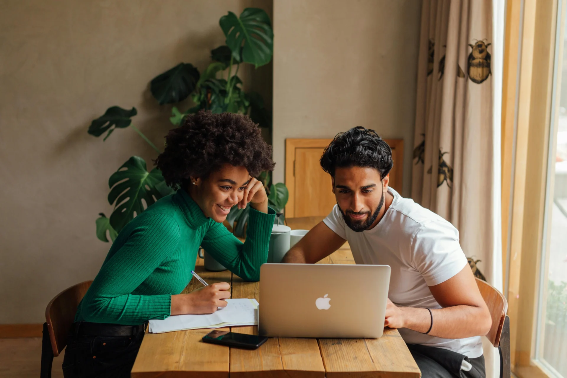 A smiling man and woman sit at a wooden table, looking at a laptop. The woman is writing on a notepad, and a large houseplant is in the background.