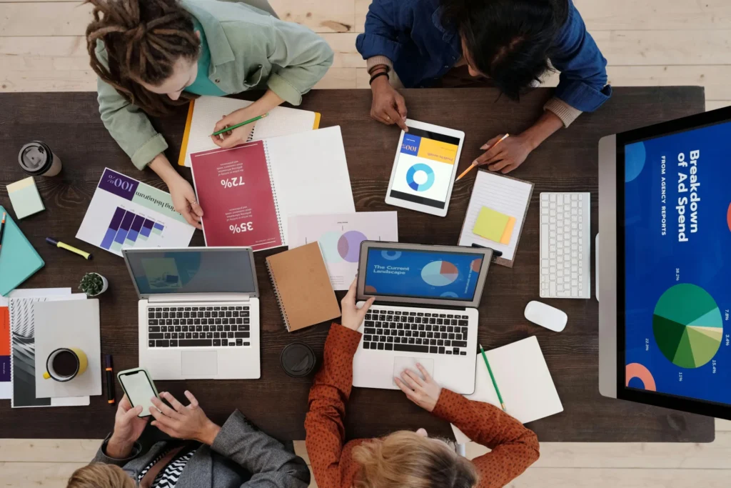 Overhead view of four diverse team members reviewing ad spend reports, charts, and laptops, planning their pillar content strategy around marketing data.