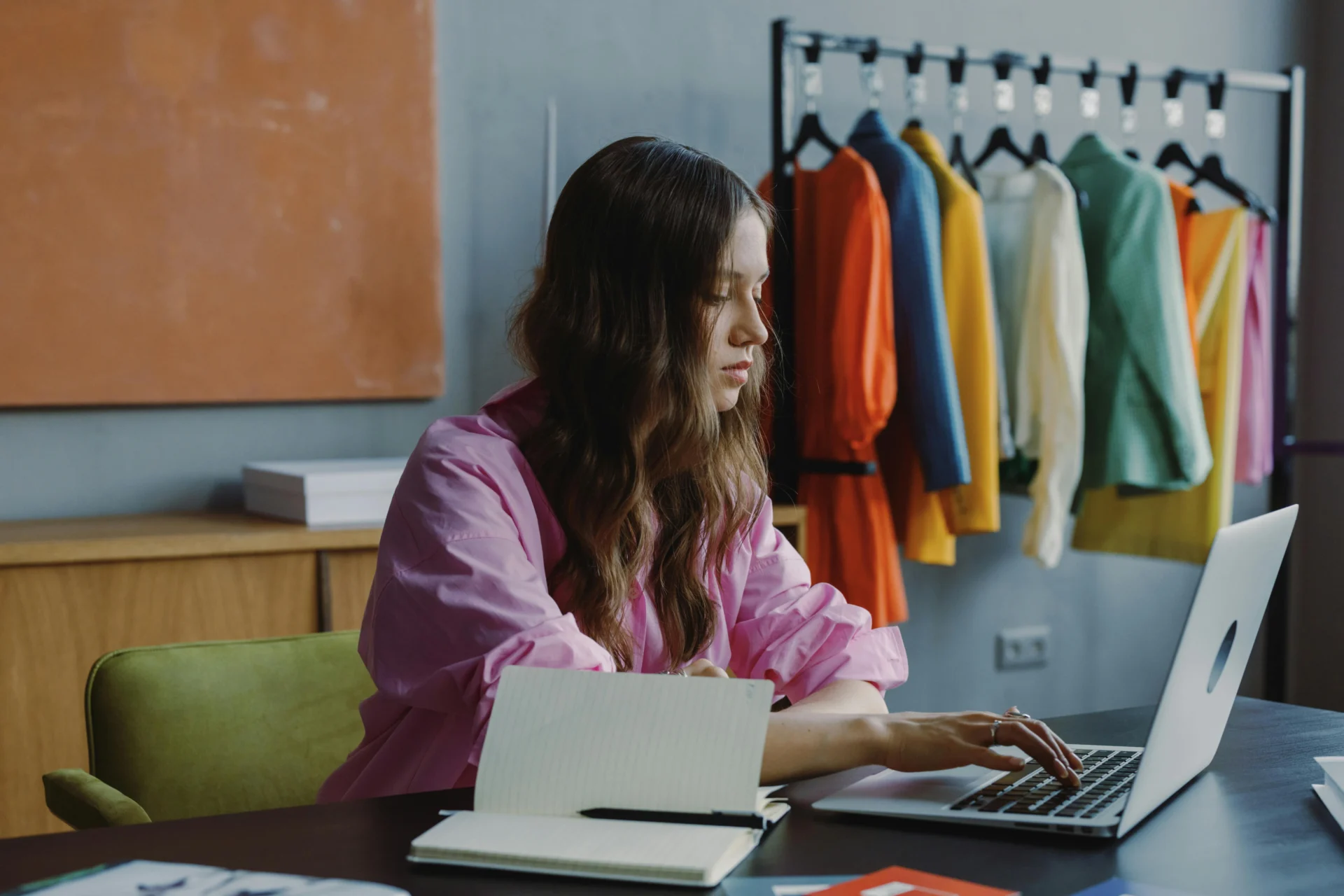 Young woman working on a laptop at a desk with colorful clothes on a rack behind her, focused on digital marketing for fashion.