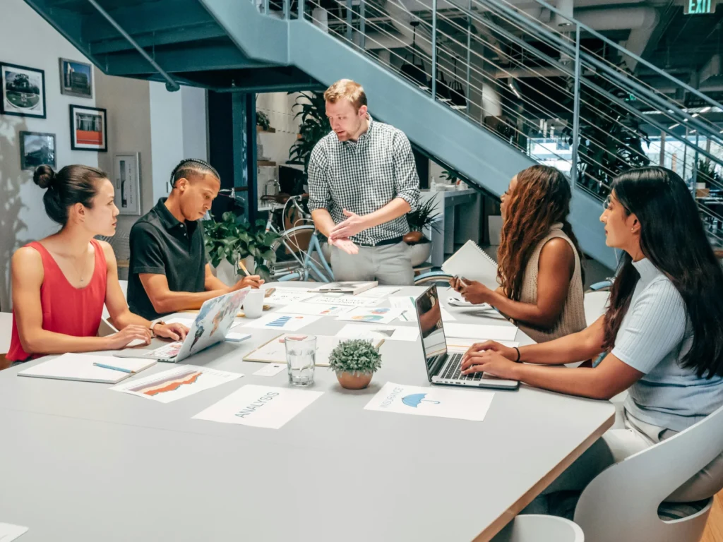 Diverse team of six professionals collaborating around a large table with laptops and charts, strategizing to generate B2B leads.