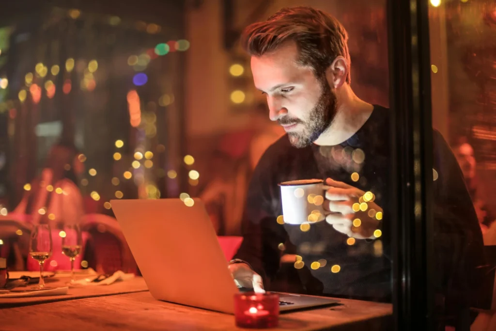 Person working on a laptop at a cafe table with fairy lights, implementing performance based marketing campaigns.
