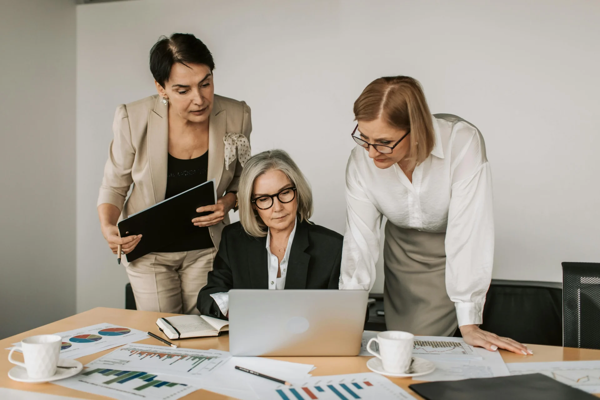 Three senior businesswomen collaborating over a laptop and printed charts, discussing the tactics for what is B2B content marketing.
