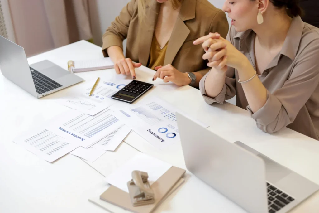 Two businesswomen reviewing financial reports and an accounting summary, calculating figures related to an evergreen content strategy budget.