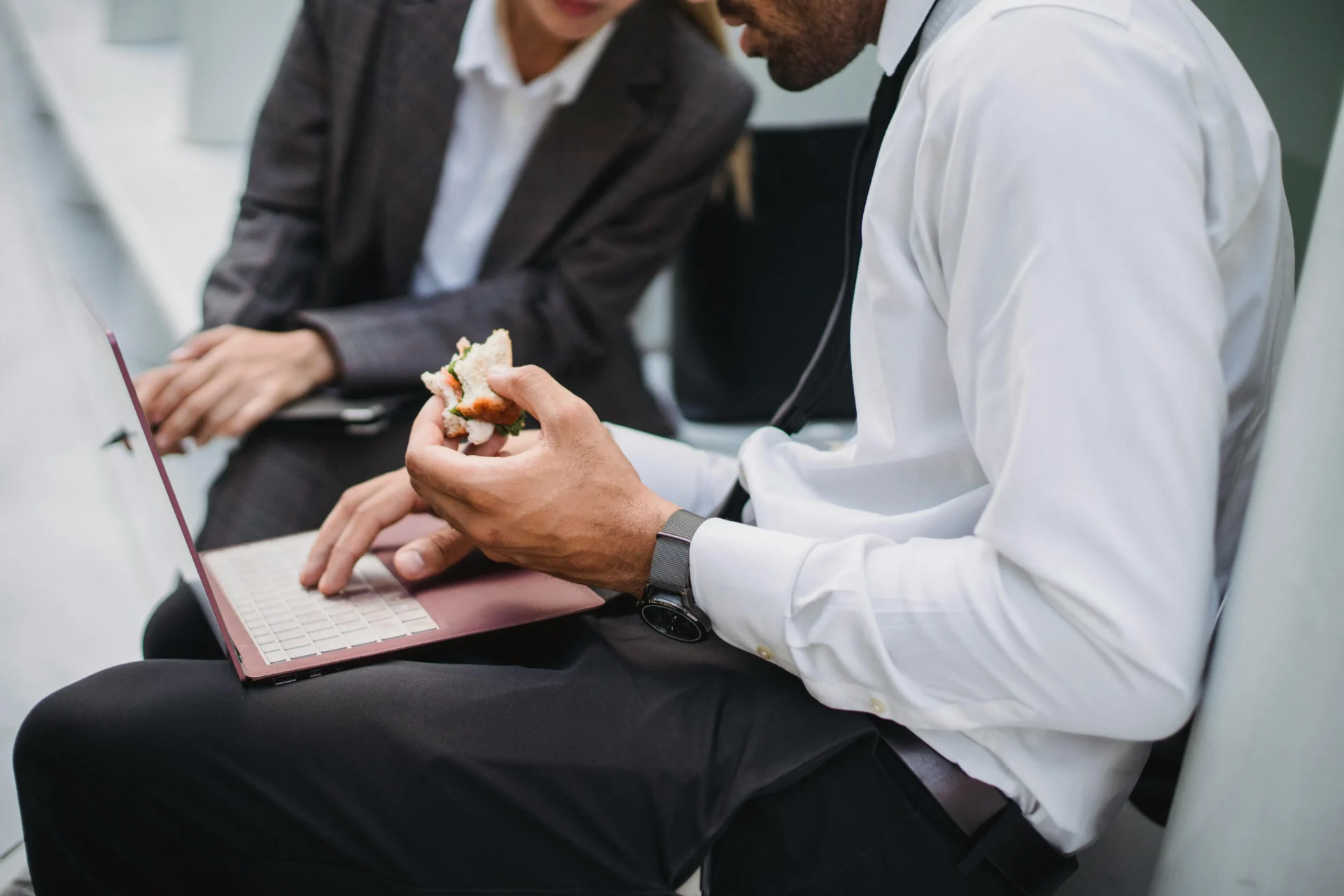 Two business professionals, one eating a sandwich while typing on a laptop, discussing the algorithm update impact on their work during a brief break.