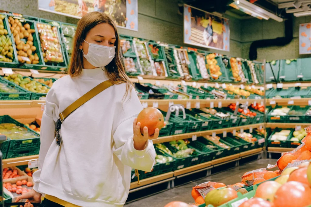 Woman in a face mask holds a mango in the produce aisle, influenced by smart grocery advertising.