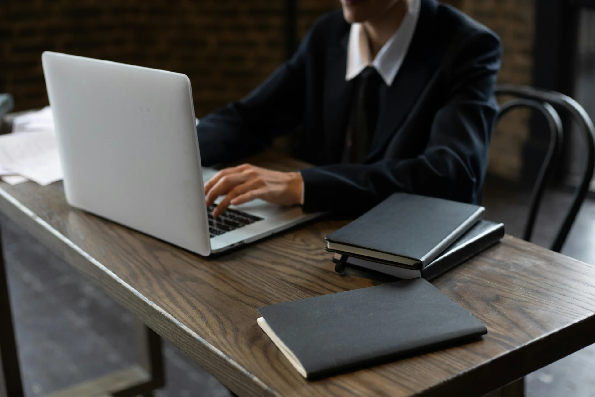 A professional in a dark suit typing on a laptop at a wooden desk with black notebooks to maintain page intent alignment for digital projects.